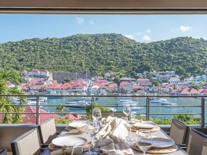A dining table set with plates, glasses, and cutlery, overlooking a marina with boats and a coastal town with red-roofed buildings.