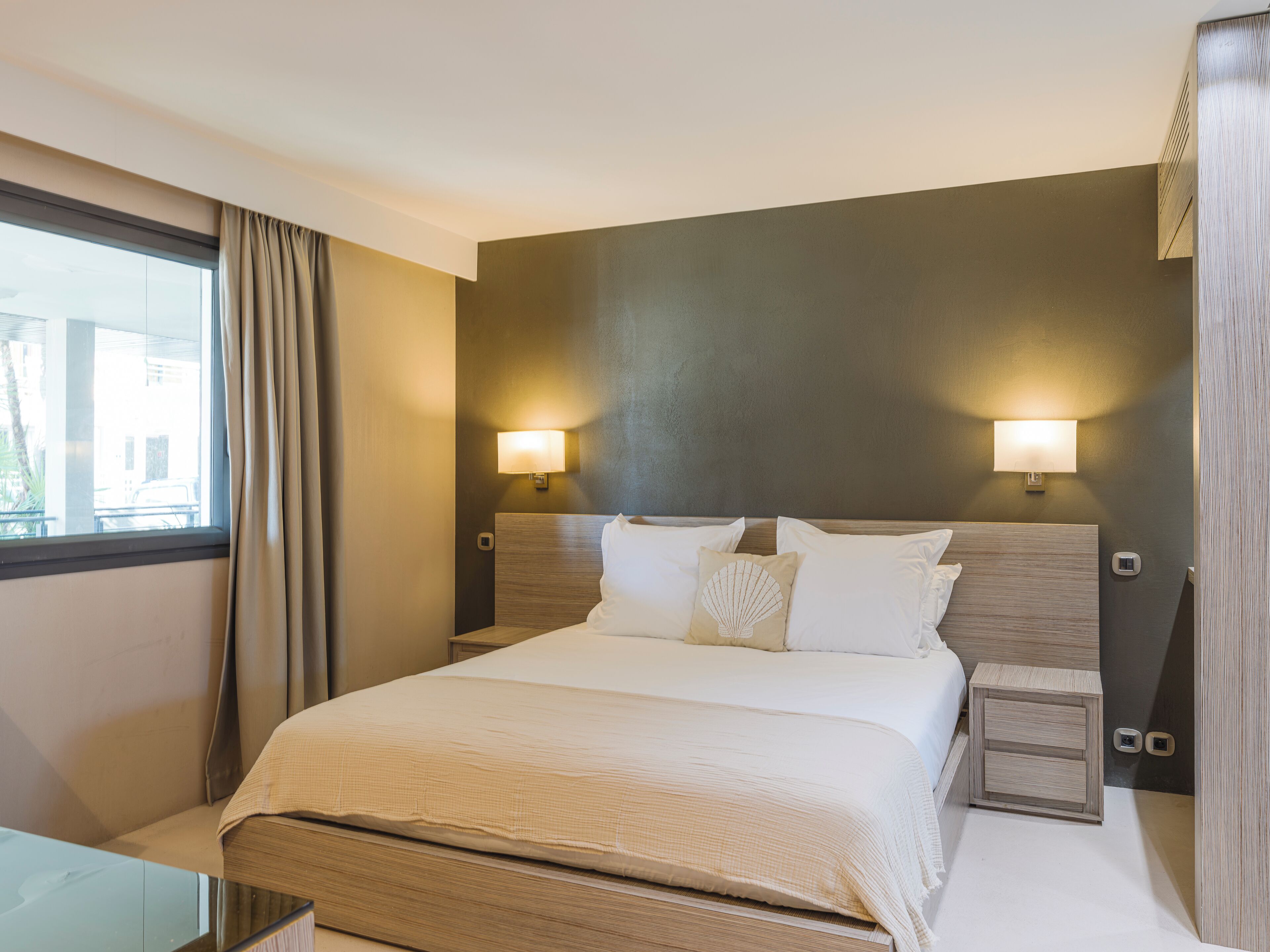 Une chambre d’hôtel moderne avec un grand lit, deux tables de chevet, une petite table, une table en verre et une fenêtre avec des rideaux.