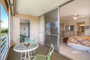 Un balcon avec une table et des chaises en verre, une porte-fenêtre coulissante donnant sur une chambre à coucher équipée d’un lit et d’une commode, et une vue sur l’extérieur.