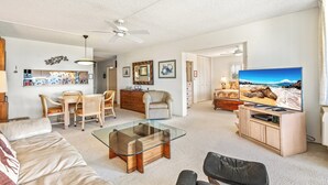 A spacious living room with a glass coffee table, a sofa, a TV, and a dining area with chairs and a table. - Kamaole Beach Royale #312 3nd Flr, Ocean View, Steps from Beach, Full Kitchen (Kihei)
