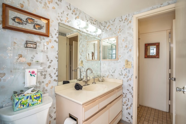 A bathroom with a large mirror, a sink, and a toilet. There is a framed picture above the mirror and a small shelf with decorative items.