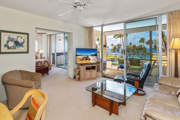 A spacious living room with a large TV, comfortable seating, and a balcony with a view of the ocean.