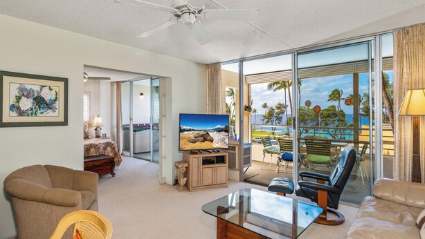 A spacious living room with a large TV, comfortable seating, and a balcony with a view of the ocean. - Kamaole Beach Royale #312 3nd Flr, Ocean View, Steps from Beach, Full Kitchen (Kihei)