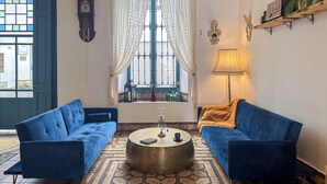 A room with two blue sofas, a round coffee table, a window with curtains, a wall clock, and a lamp. - La Tienda Nueva (Còmpeta)