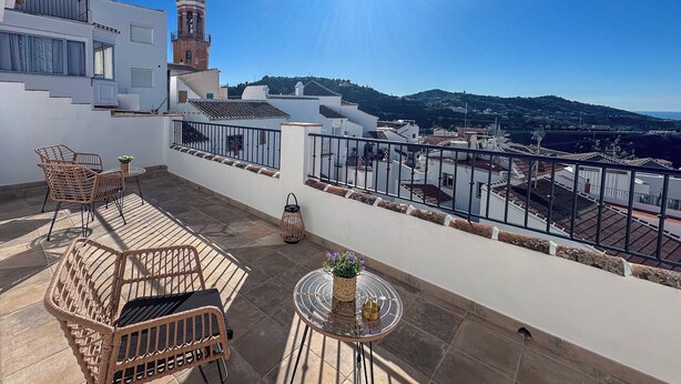A terrace with wicker furniture, a small round table, and a view of a town with buildings and a hill in the distance. - La Tienda Nueva (Còmpeta)