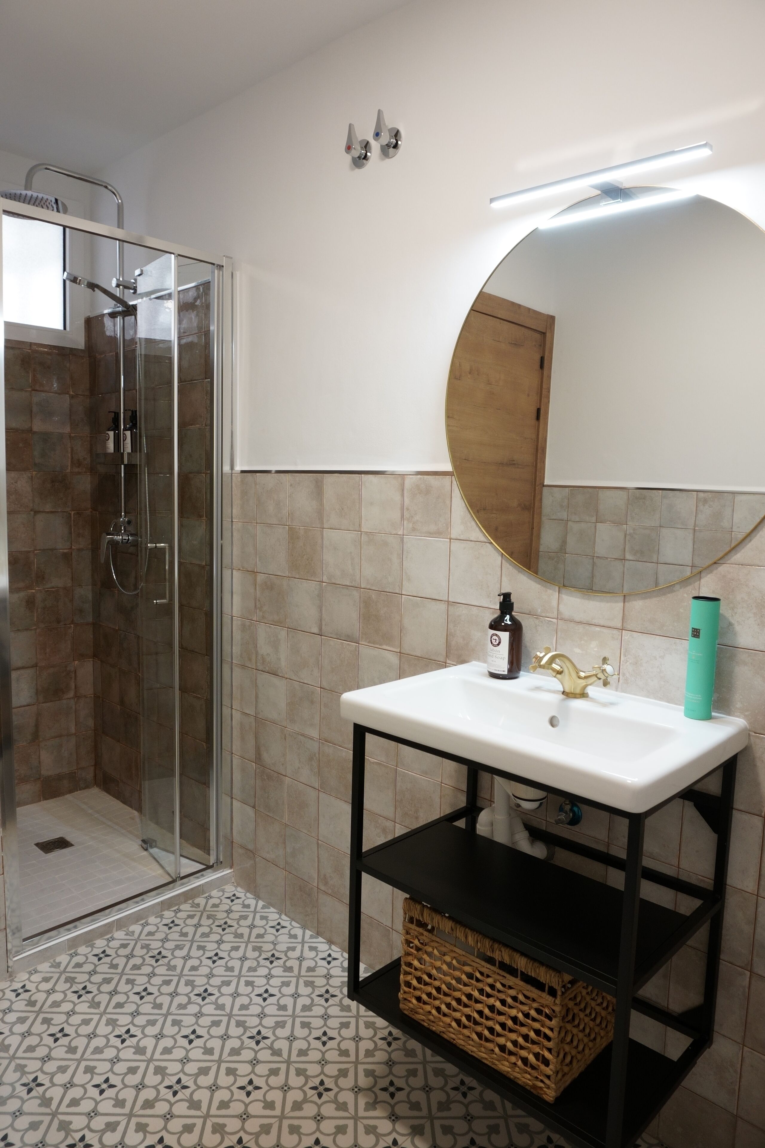 Een badkamer met een douche met glazen wand, een witte wastafel met een gouden kraan, en een ronde spiegel erboven.