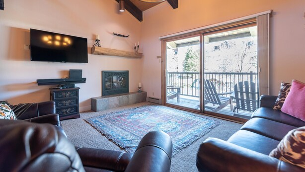 A living room with a large flat-screen TV, a fireplace, a large window, and a balcony with outdoor seating. - Large Deck + Fireplace Hot Tubs- Walk to Mtn (Steamboat Springs)