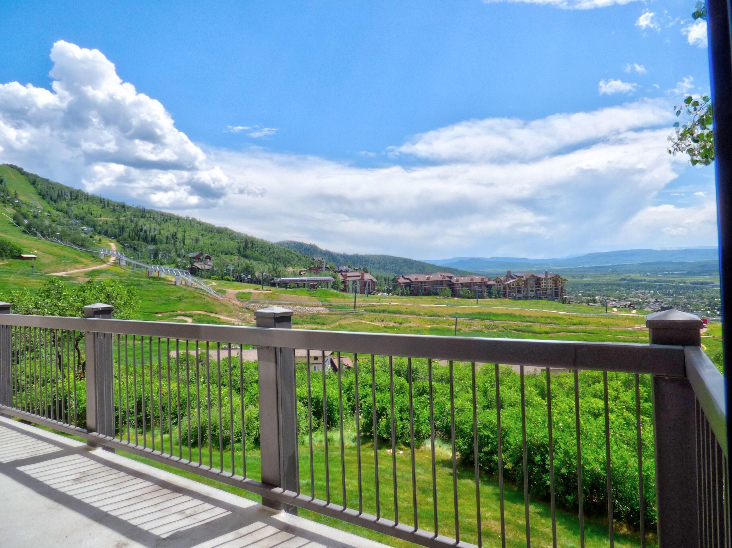 A balcony with a view of a green landscape, a railway track, and a building complex.