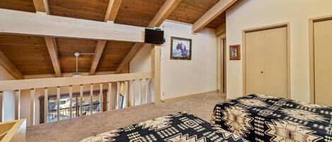 A bedroom with a sloped ceiling, a ceiling fan, a bed with patterned bedding, a television, and a door.