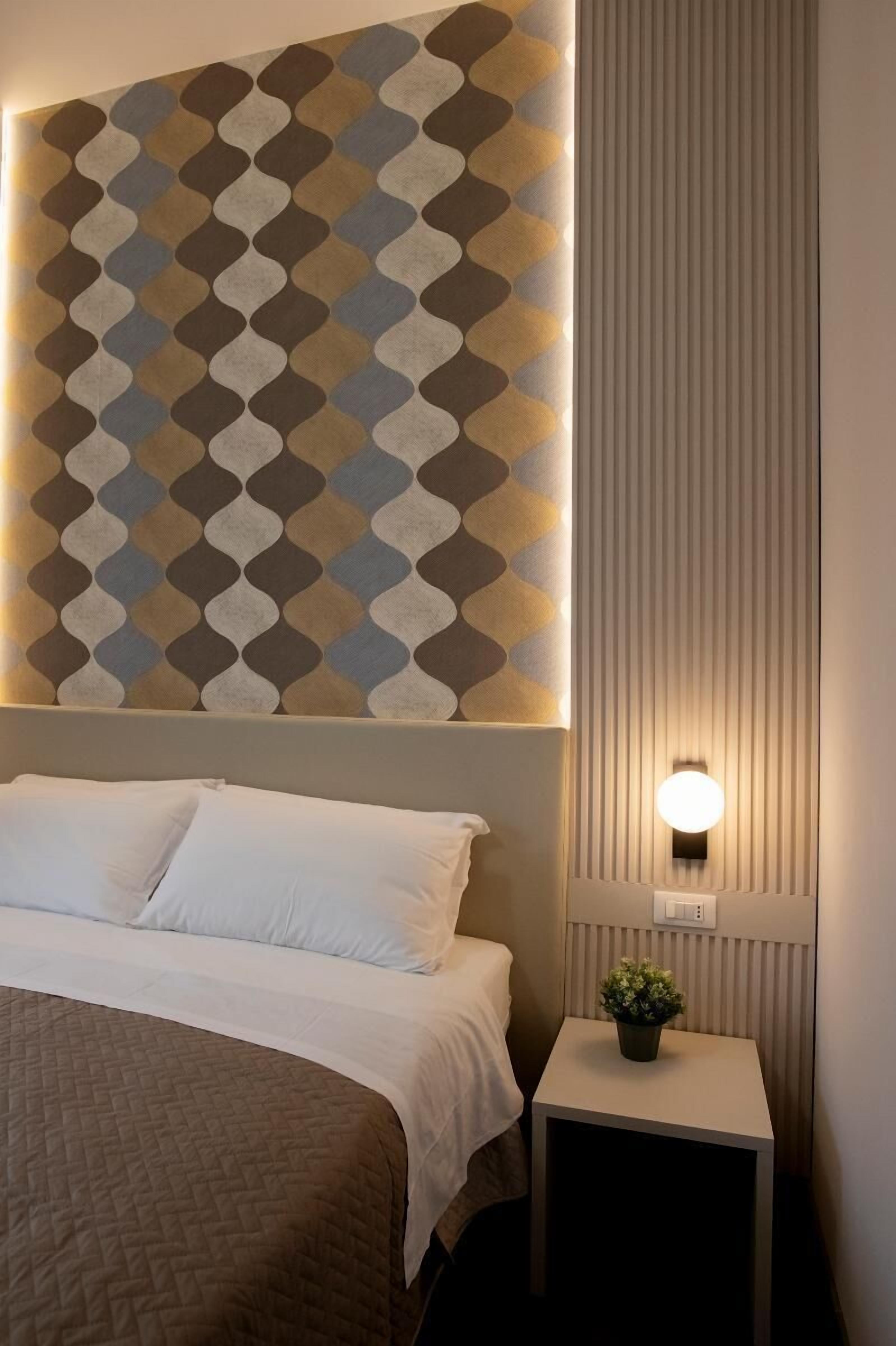 A neatly made bed with a quilted cover, a wall with a geometric patterned wallpaper, a small bedside table with a potted plant, and a wall-mounted light fixture.