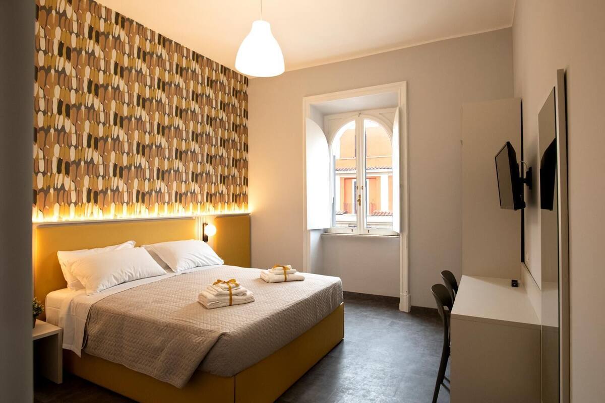 Ein modernes Hotelzimmer mit einem großen Bett, einem gemusterten Kopfteil, einem Nachttisch mit Lampe, einem Fenster mit bogenförmigem Design und einem an der Wand montierten Flachbildfernseher.