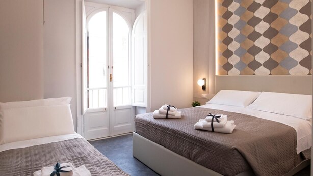 A hotel room with two beds, a large window, a patterned wall, and a bedside table with a lamp. - DoppioSei -Guest House (Rome)
