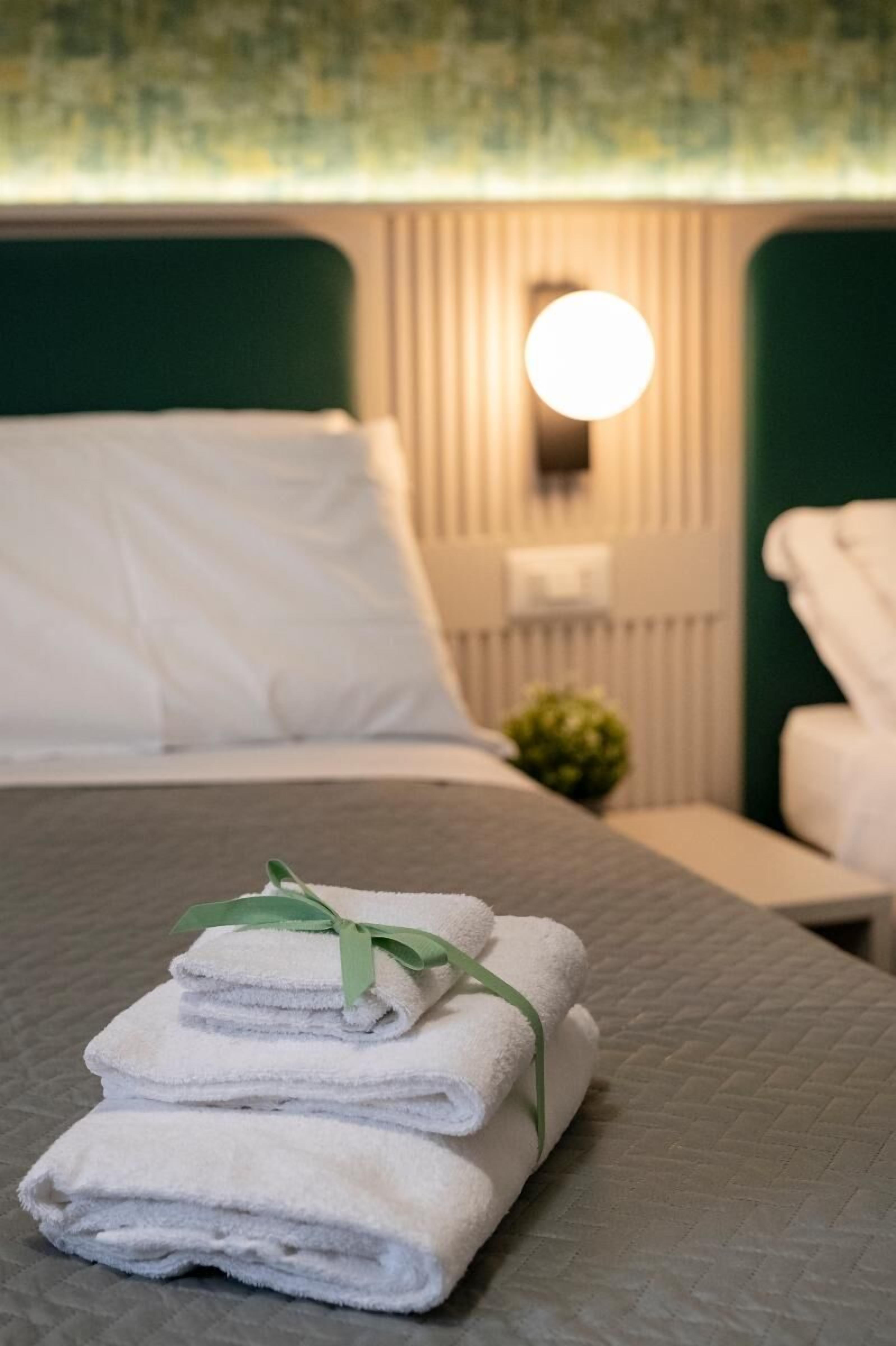 Ein ordentlich bezogenes Bett mit gefalteten weißen Handtüchern und einem grünen Band, eine Nachttischlampe und eine kleine Pflanze auf einem Nachttisch.