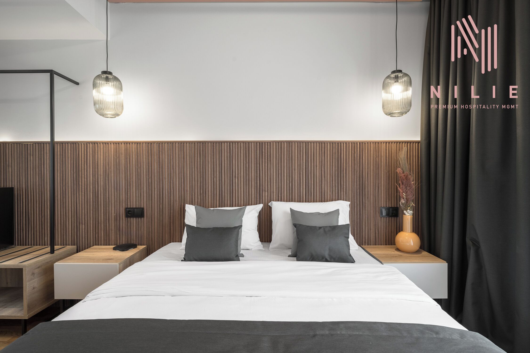 A modern hotel room with a large bed, two bedside tables, a TV, and pendant lighting.