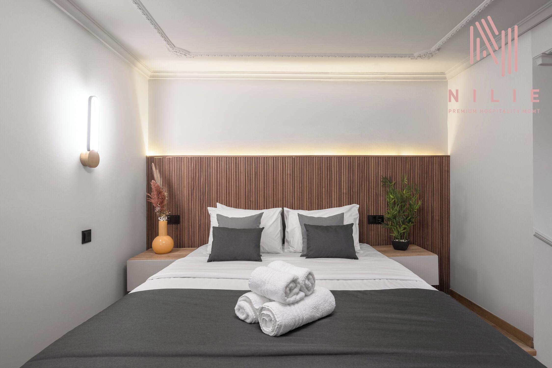 A modern hotel room with a neatly made bed, a bedside table with a vase, and a wall-mounted light fixture.