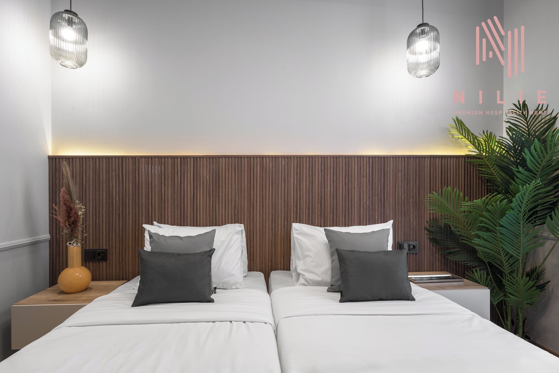 Moderni hotellihuone, jossa on kaksi sänkyä, puinen pääty, riippuvalaisimet, ruukkukasvi ja maljakko kuivakukilla.