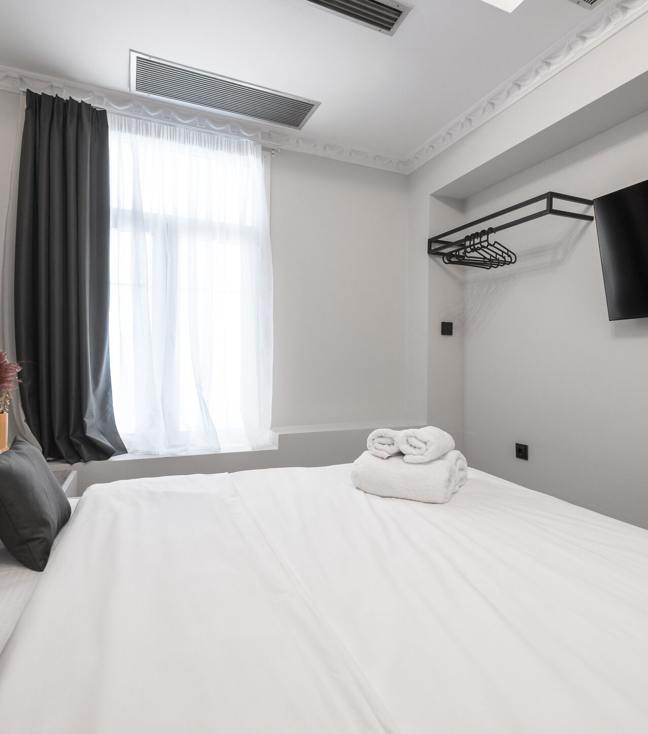 A hotel room with a neatly made bed, a window with curtains, a wall-mounted TV, and a clothes drying rack.