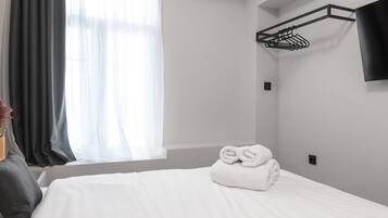 A hotel room with a neatly made bed, a window with curtains, a wall-mounted TV, and a clothes drying rack.