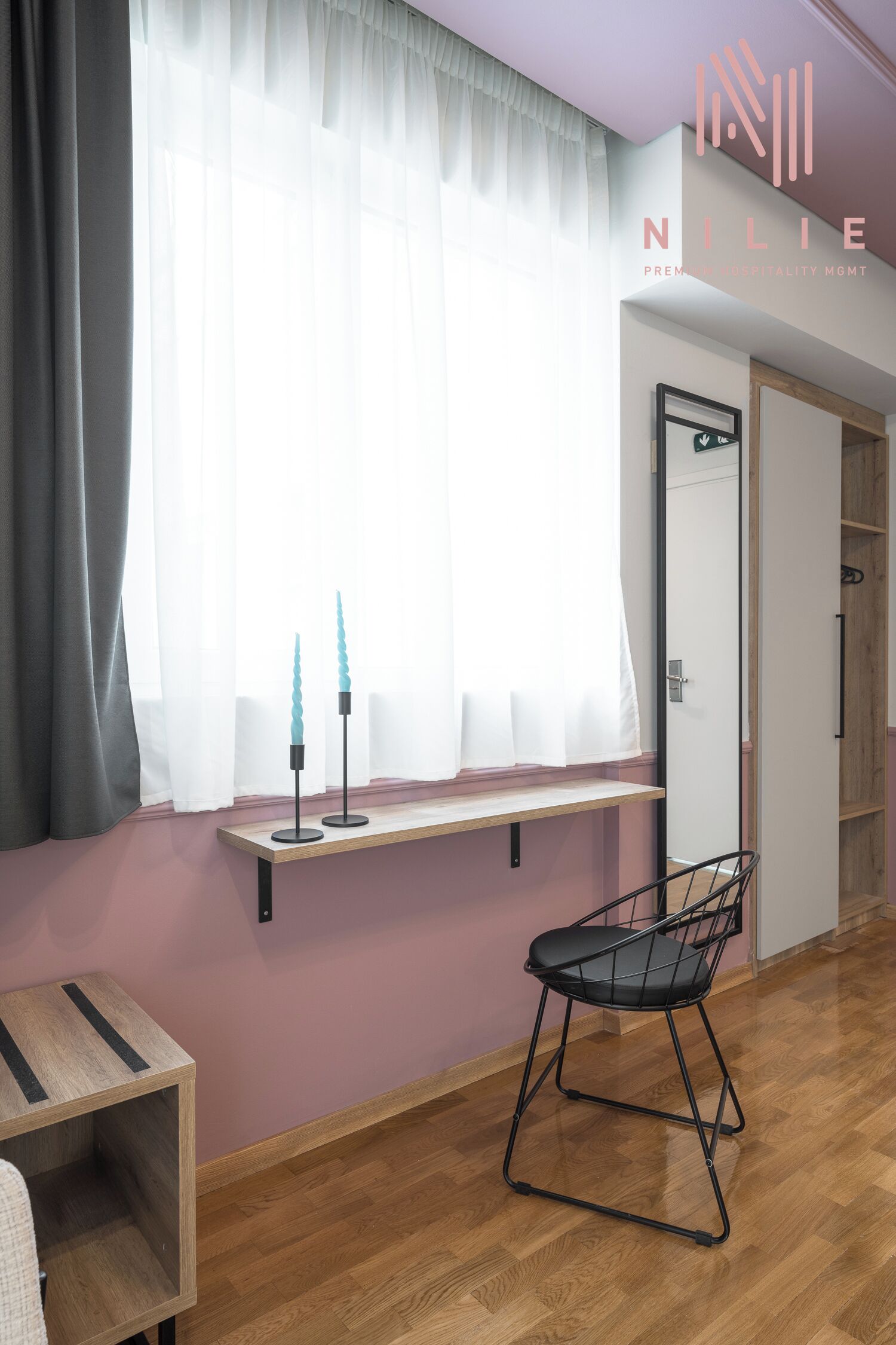 A room with a wooden floor, a black chair, a small table, a window with sheer curtains, and a shelf with two teal candles.