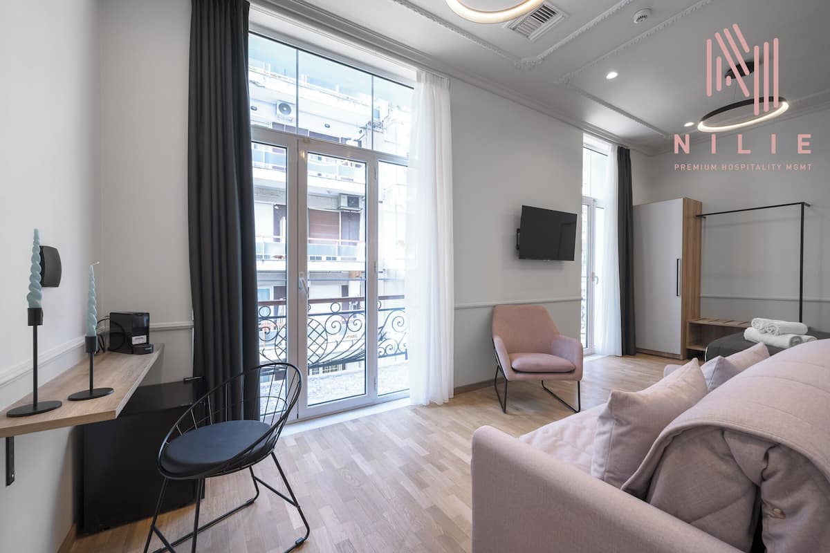 A modern hotel room with a balcony, a desk with a chair, a sofa, and a TV mounted on the wall.