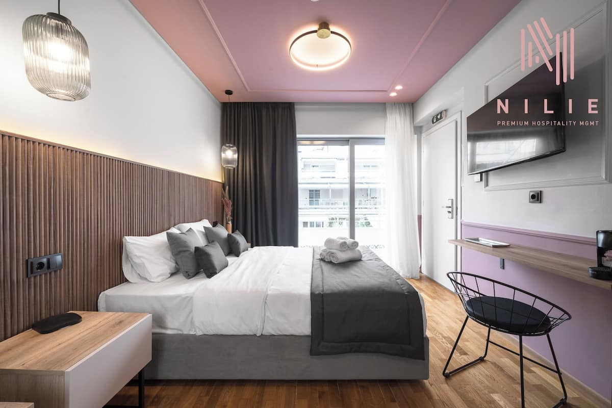 A modern hotel room with a bed, a desk, a chair, a TV, and a view of the city.