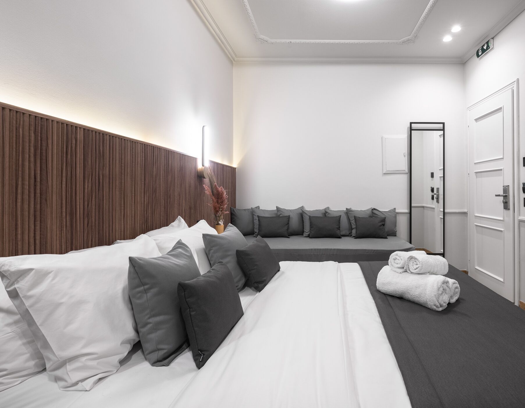 Une chambre d’hôtel moderne avec un grand lit, une literie aux tons de gris et de blanc, une tête de lit en bois, un coin salon avec des coussins et une porte dotée d’un miroir.