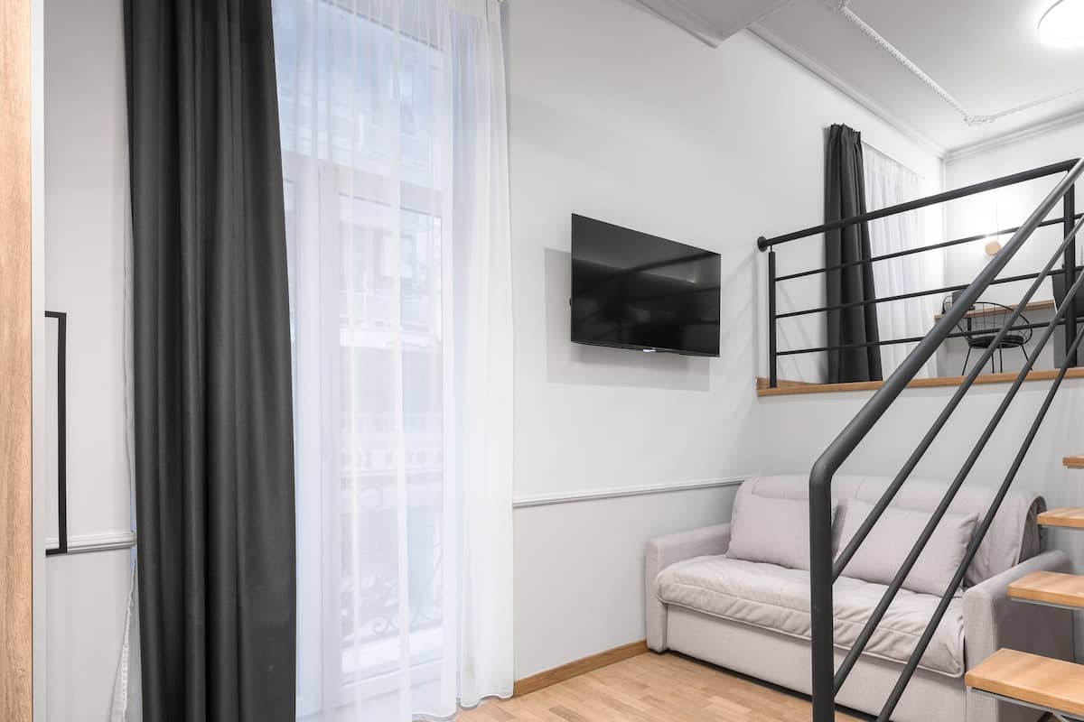 A modern living room with a sofa, a TV mounted on the wall, a staircase with a railing, and a large window with sheer curtains.