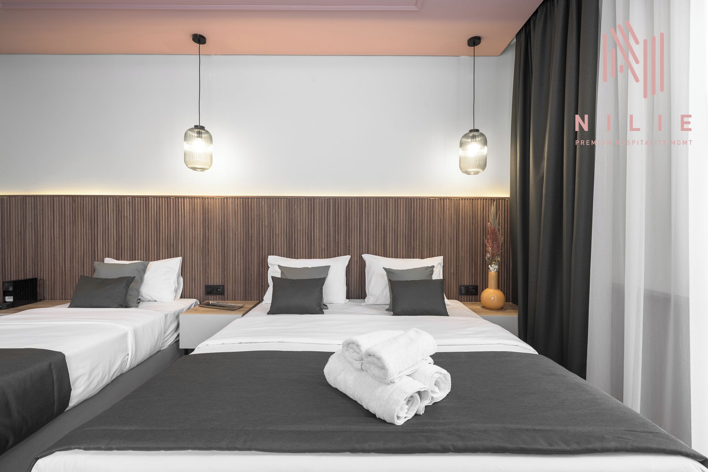 Una habitación de hotel moderna con dos camas, un ventanal con cortinas y una lámpara fijada a la pared.