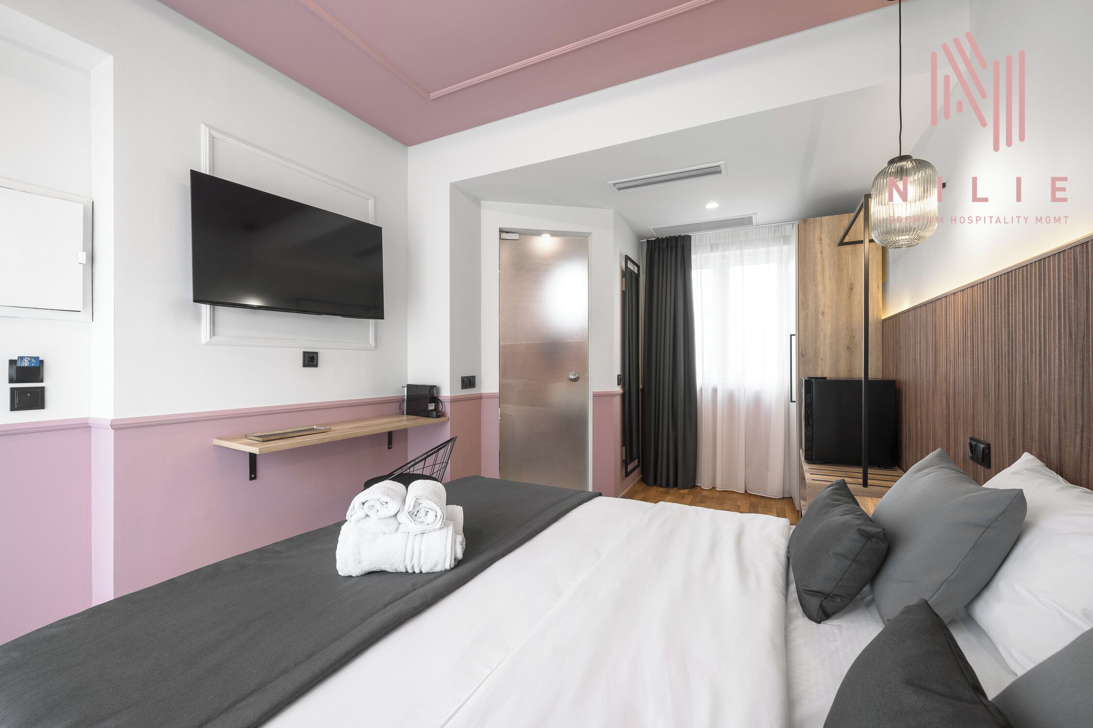 Una habitación de hotel moderna con una cama, un televisor montado en la pared, una mesita de noche y una lámpara colgante.