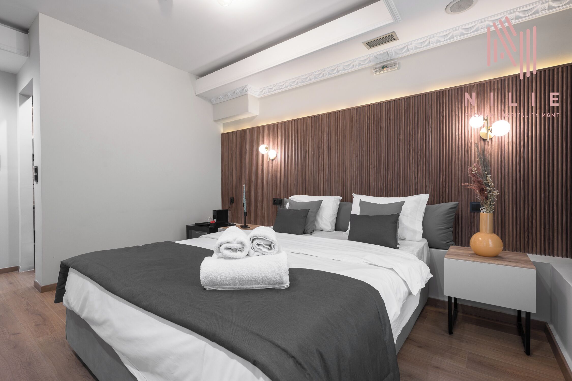 Een moderne hotelkamer met een groot bed, een nachtkastje, een houten accentmuur en een vaas met gedroogde bloemen.