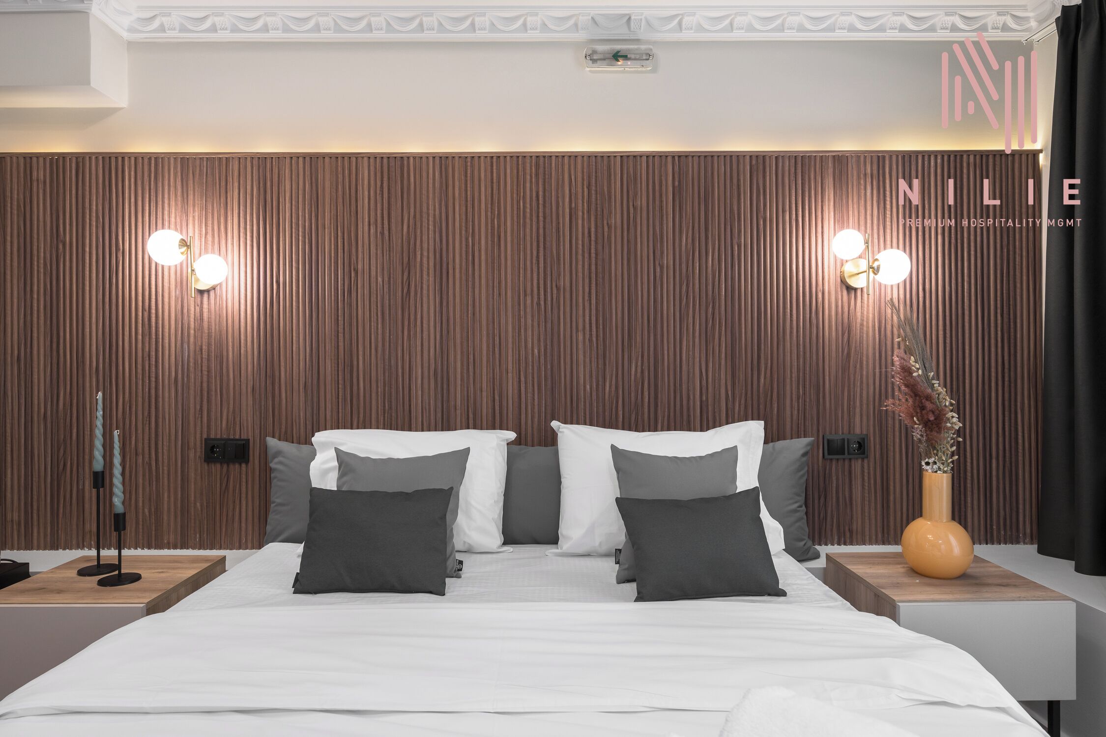 Et moderne hotelværelse med en træsenggavl, en stor seng med hvid og grå sengetøj, to sengeborde med lamper, og en vase med tørrede blomster på højre side.