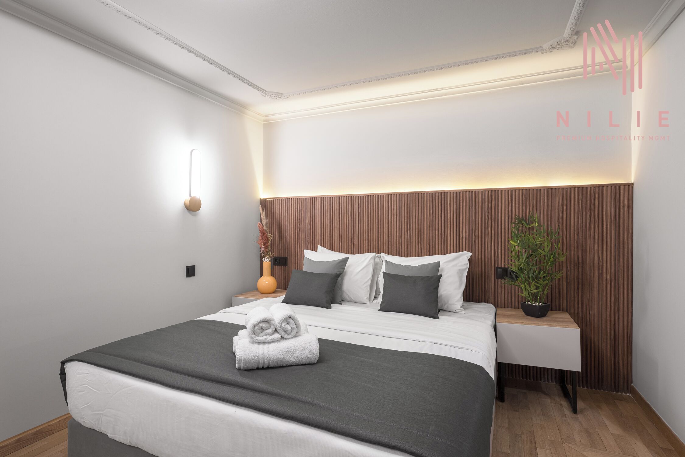 A modern hotel room with a neatly made bed, a bedside table with a plant, and a wall-mounted light fixture.