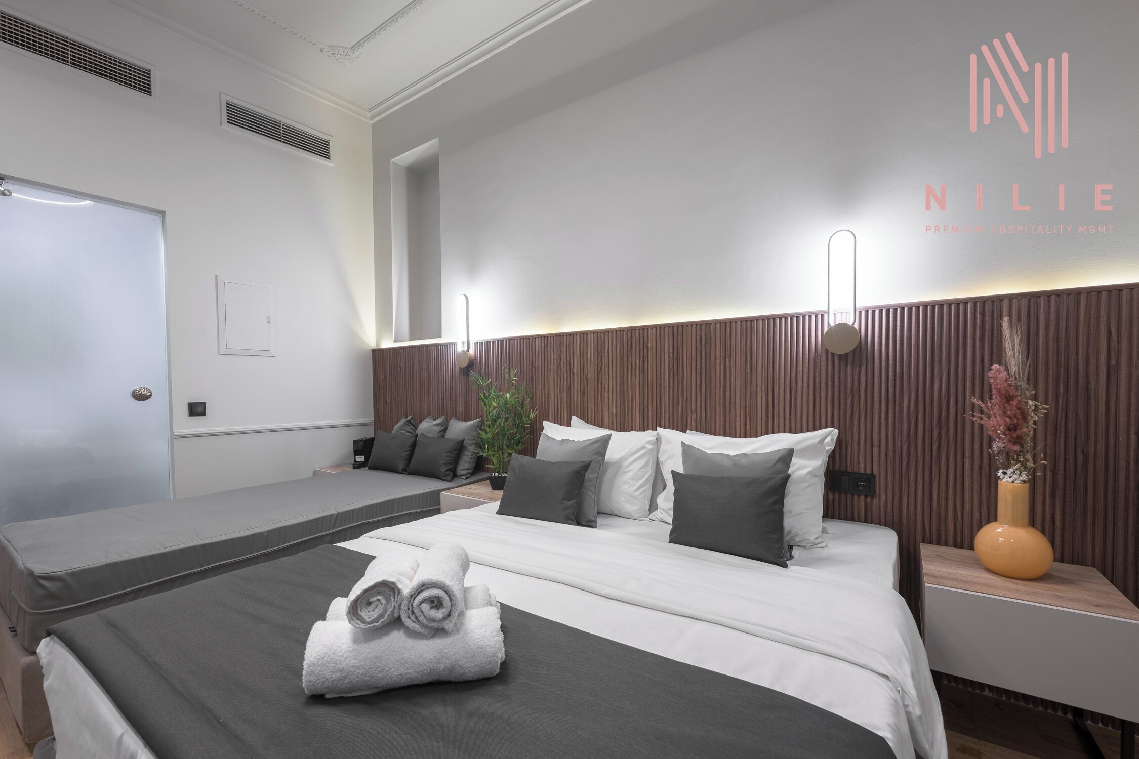 Una habitación de hotel moderna con una cama grande, un sofá cama más pequeño, una mesita de noche con un florero y una lámpara de pared.