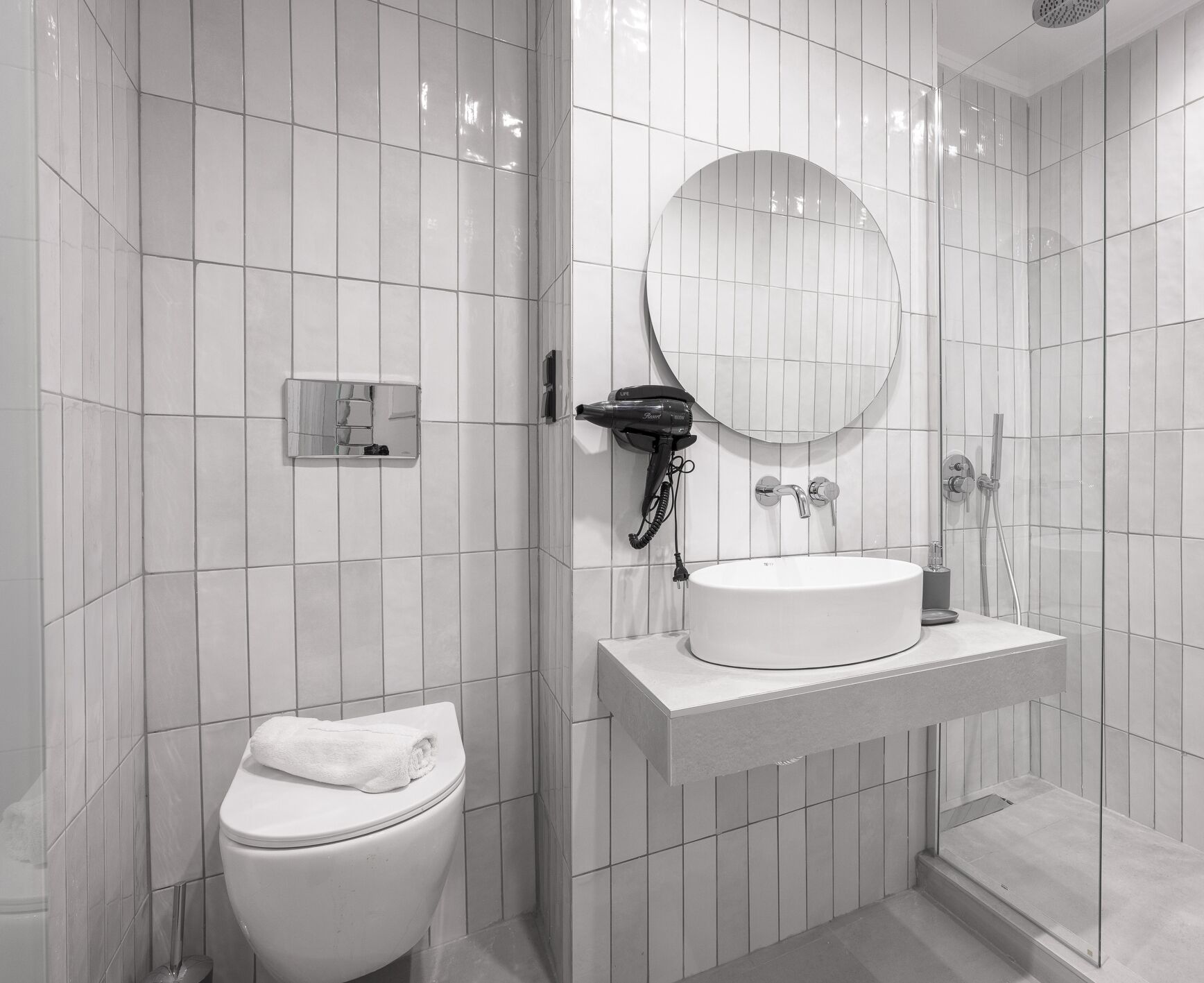 A modern bathroom with white tiles, a round mirror, a hairdryer, a sink, and a toilet.