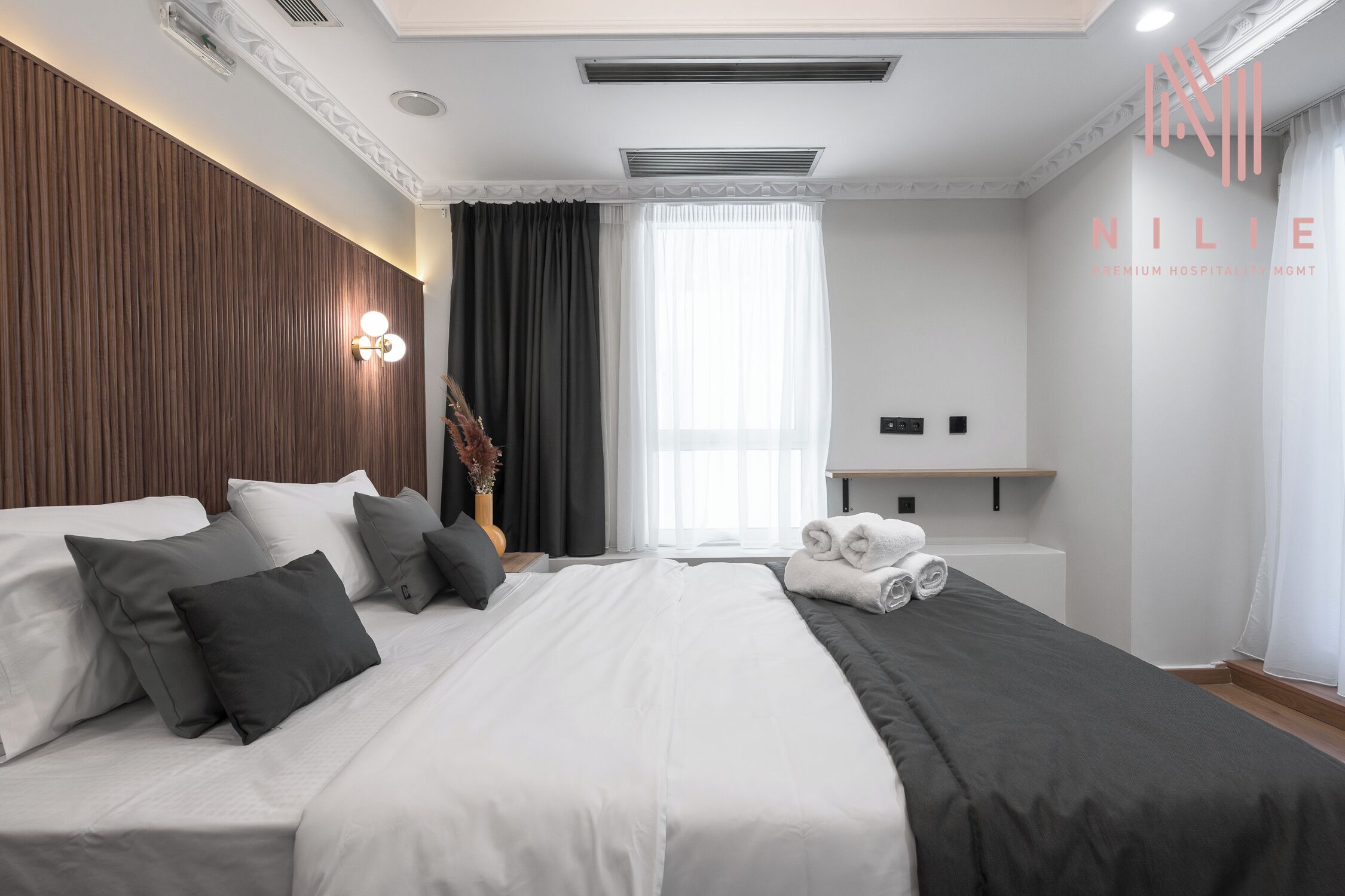 A modern hotel room with a large bed, a flat-screen TV, a wooden headboard, and a large window with curtains.