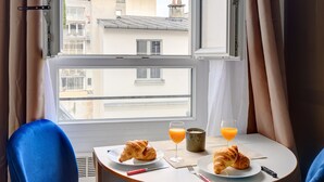 Una habitación con una mesa redonda dispuesta para el desayuno, dos sillas azules, una ventana con cortinas y vistas a edificios.