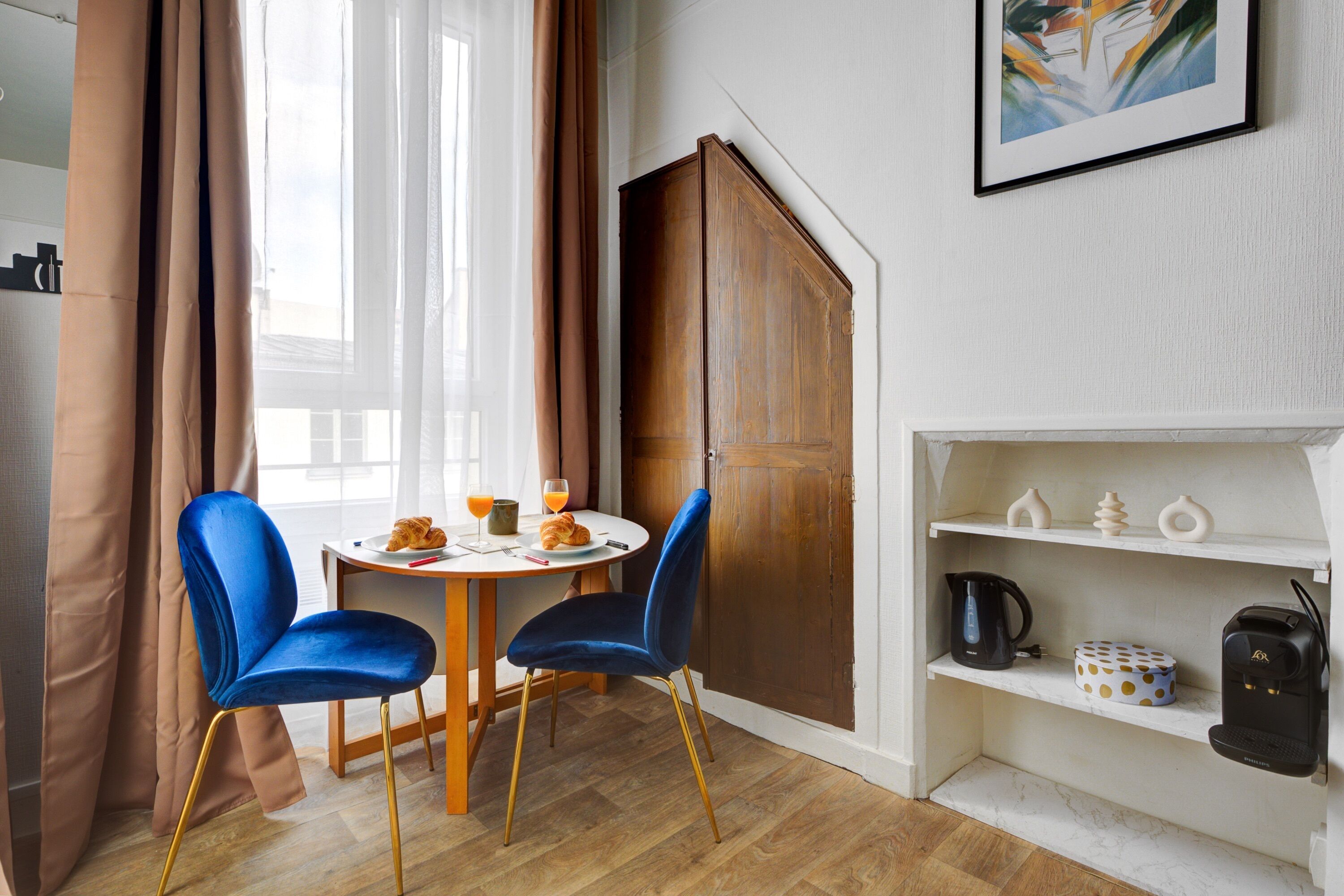 A compact dining area with two blue chairs, a small round table, and a coffee machine on a white shelf.