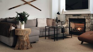 A modern living room with a stone fireplace, a grey sofa, a coffee table, and a TV mounted above the fireplace. - Halte MB2 — Terrace, Pool, Lake & Hiking (Mont-Blanc)