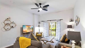 Een woonkamer met een bank, fauteuil, salontafel, televisie, plafondventilator en een raam met gordijnen.