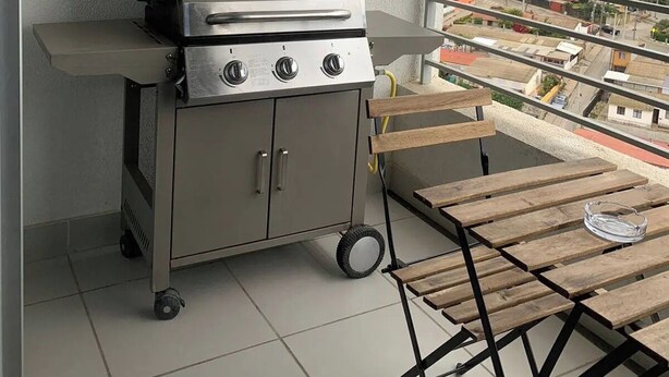 Un balcon équipé d’un barbecue mobile, d’une table en bois et offrant une vue sur le paysage urbain.