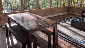 Un intérieur en bois avec une grande table et des bancs, de grandes fenêtres donnant sur la forêt et un ventilateur de plafond.