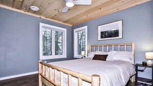 Une chambre à coucher avec un lit en bois, deux fenêtres, un ventilateur de plafond et un tableau encadré au mur.