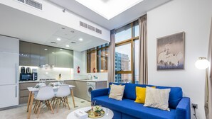 Ein modernes Wohnzimmer mit einem blauen Sofa, einem Esstisch mit Stühlen, einer Küchenzeile und einem großen Fenster mit Vorhängen.