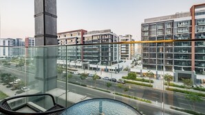 Ein modernes Gebäude mit Glasfassade, einem Balkon mit Glasgeländer und Blick auf eine Straße mit Autos und Palmen.