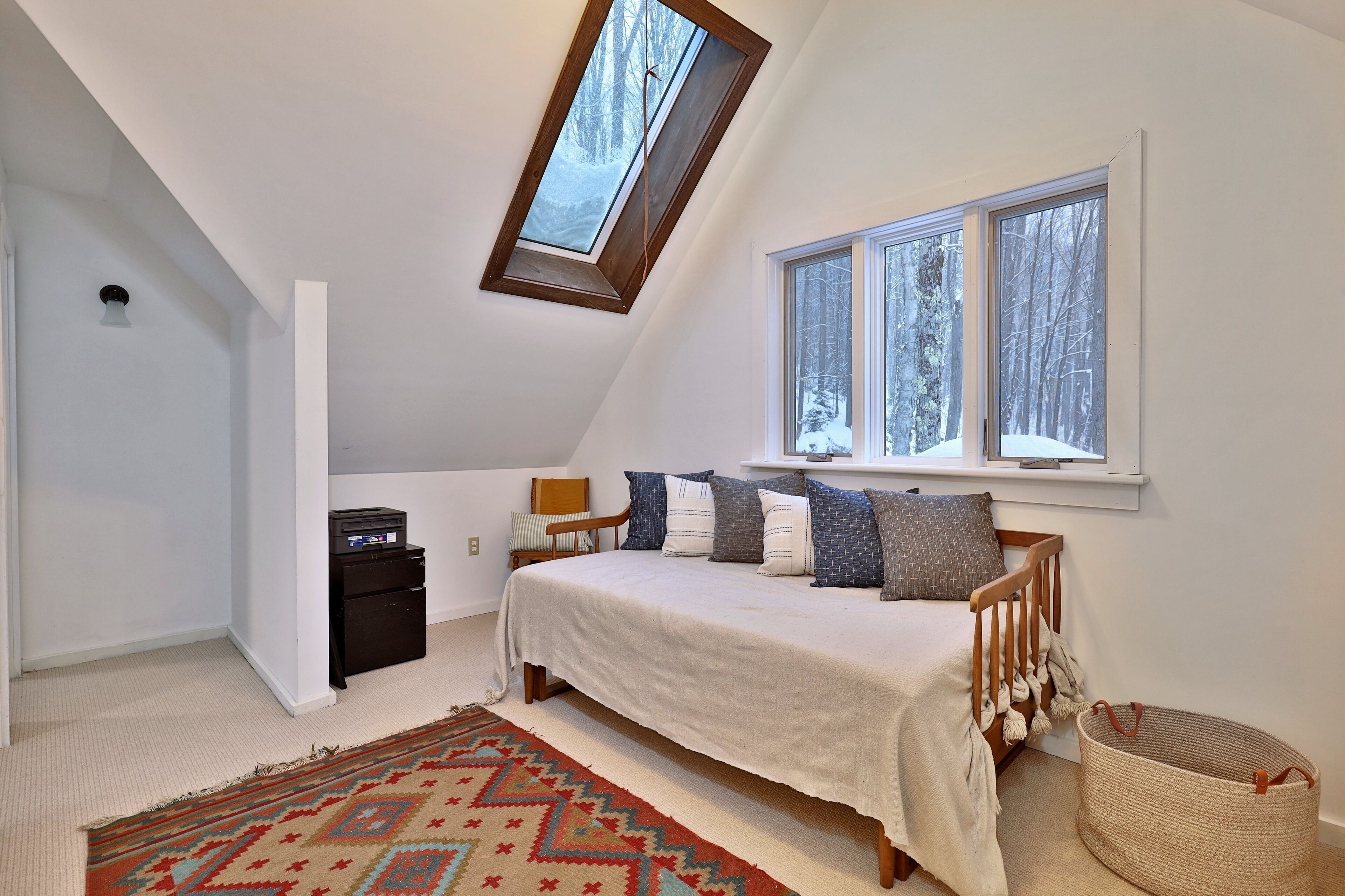 A bedroom with a sloped ceiling, a skylight, a bed with pillows, a nightstand, a lamp, and a window with a snowy view.