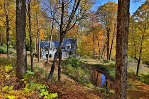 A rustic cabin surrounded by autumn foliage, a small stream, and a wooden deck. - Mountainview Escape 4 Bedroom Home (Killington)