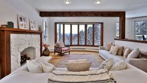 A cozy living room with a fireplace, a large window with a snowy view, a sofa set, and a wooden ceiling. - Mountainview Escape 4 Bedroom Home (Killington)