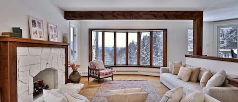 A cozy living room with a fireplace, a large window with a snowy view, a sofa set, and a wooden ceiling.