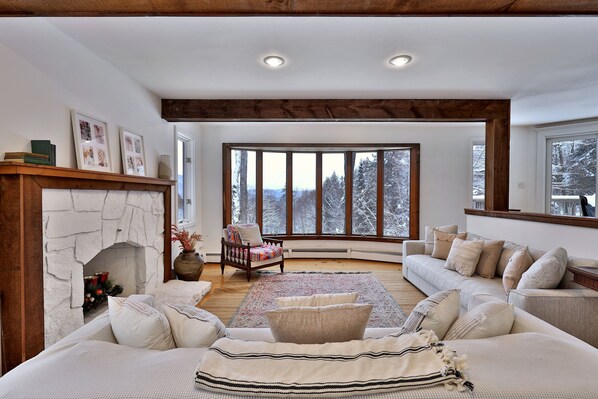 A cozy living room with a fireplace, a large window with a snowy view, a sofa set, and a wooden ceiling. - Mountainview Escape 4 Bedroom Home (Killington)