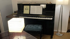 A piano room with a piano, sheet music, a framed picture, a bench, and a lamp.